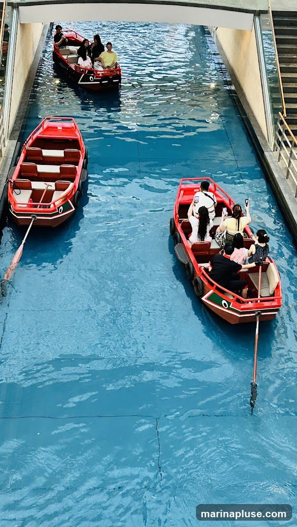 Several red boats are navigating a bright blue canal. Some boats are occupied by people, while others are empty. The canal appears to be indoors, possibly in a shopping mall or entertainment venue, with a walkway and stairs visible on the right side.