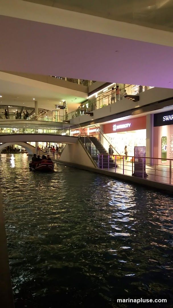 An indoor canal with a boat carrying passengers, surrounded by multi-level shops and walkways. The shops have illuminated signs and displays, with people browsing on the upper levels. The water reflects the lights from the shops and ceiling.
