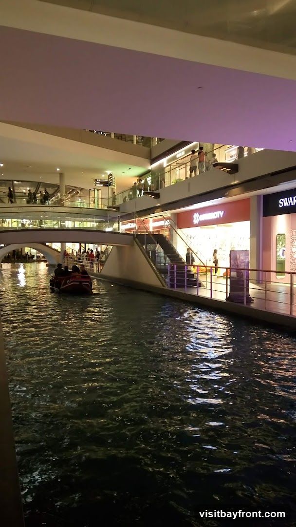 An indoor canal with a boat carrying passengers, surrounded by multi-level shops and walkways. The shops have illuminated signs and displays, with people browsing on the upper levels. The water reflects the lights from the shops and ceiling.