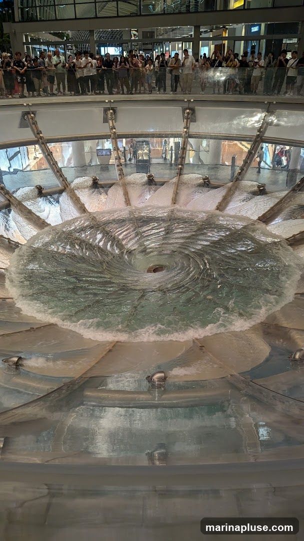 A large indoor fountain creates a vortex of water in the center, with water cascading down from multiple points around the perimeter. A crowd of people is gathered on an upper level, observing the fountain. The architecture suggests a modern shopping mall or public space.