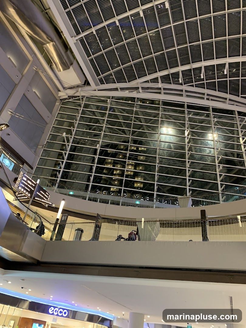 The image shows the interior of a large, modern shopping mall with a high, arched glass ceiling. Multiple levels are visible, with escalators and walkways connecting them. People can be seen browsing and walking around. A sign for 'ECCO' is visible on one of the lower levels. The overall impression is one of a spacious and well-lit commercial environment.