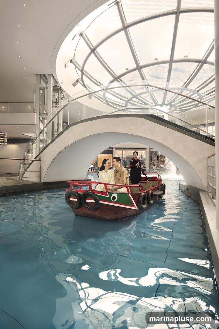 A couple is enjoying a boat ride on a canal-like water feature inside a modern building with a large glass dome ceiling. A staff member in uniform is steering the boat. The architecture features curved walkways and escalators.