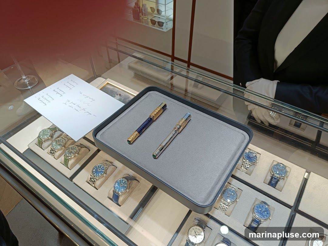 A display case filled with luxury watches and two ornate pens. A person in a suit and white gloves stands behind the counter. A handwritten note is placed on the display case.