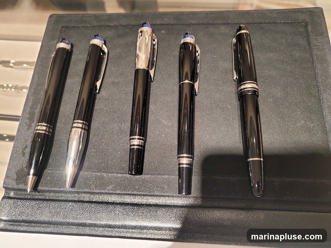 A display of five black Montblanc pens with silver accents and blue caps, arranged on a dark textured surface.