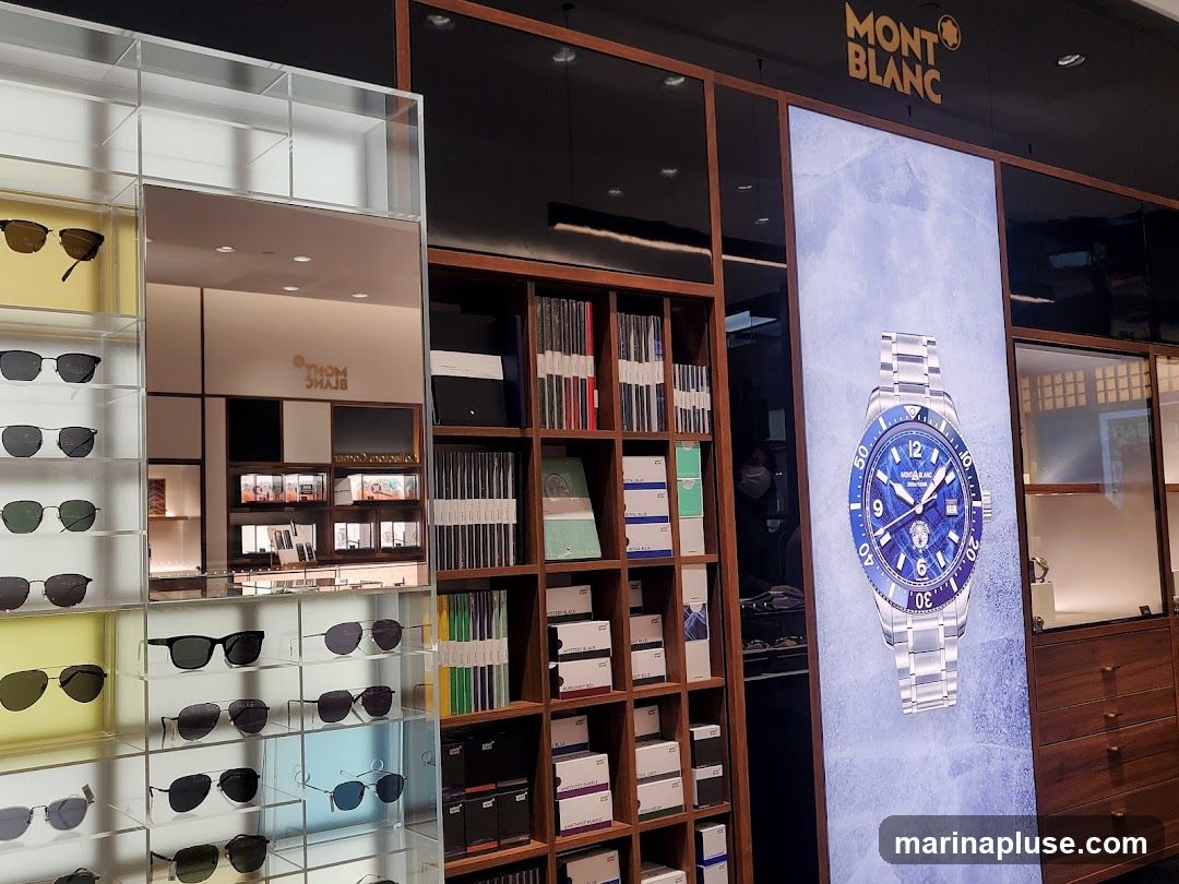 A display inside a Montblanc store featuring sunglasses on the left, shelves of products and books in the center, and a large digital screen showcasing a Montblanc watch on the right. The Montblanc logo is visible above the screen.