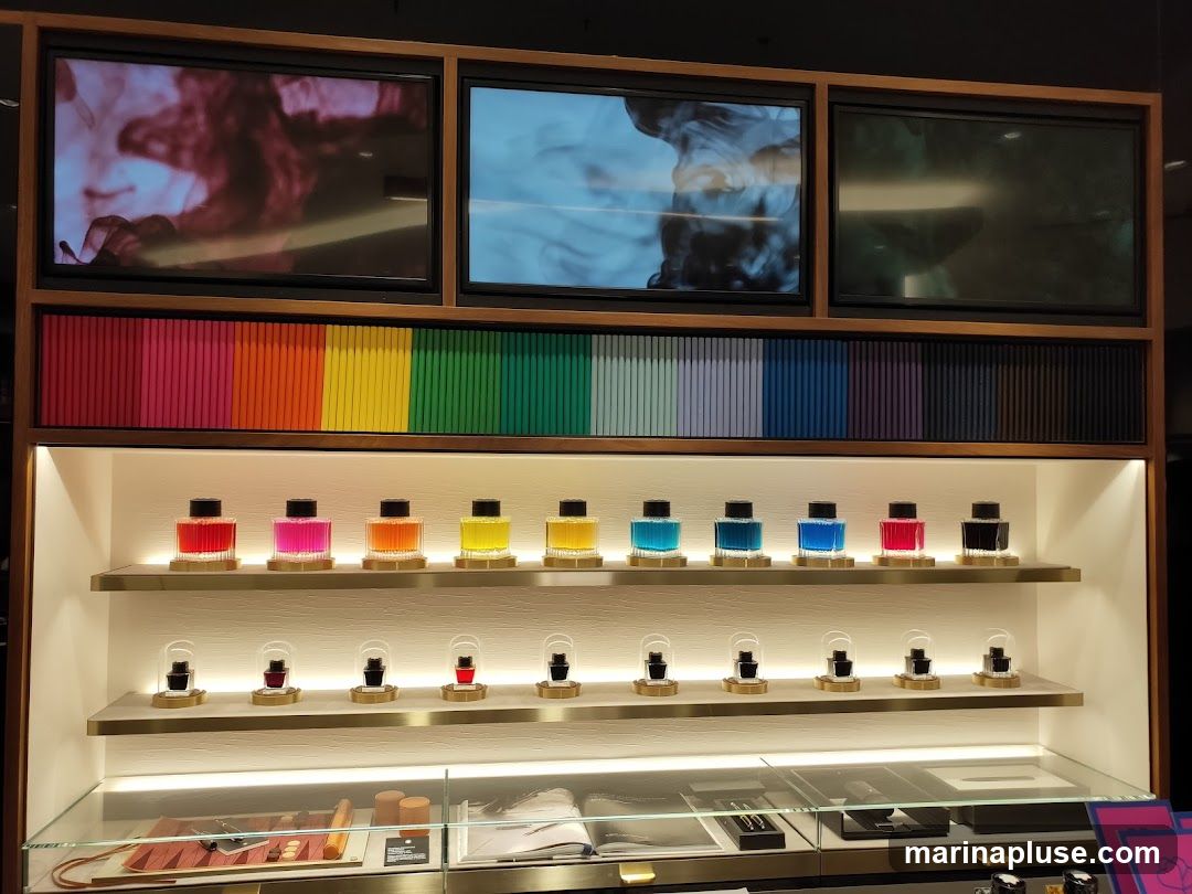 A display shelf with rows of colorful ink bottles. Above the ink bottles, there is a row of colorful spines, possibly books or ink sample cards. Three screens are mounted above the spines, showing abstract colorful patterns. The bottom shelf displays various items in glass cases.