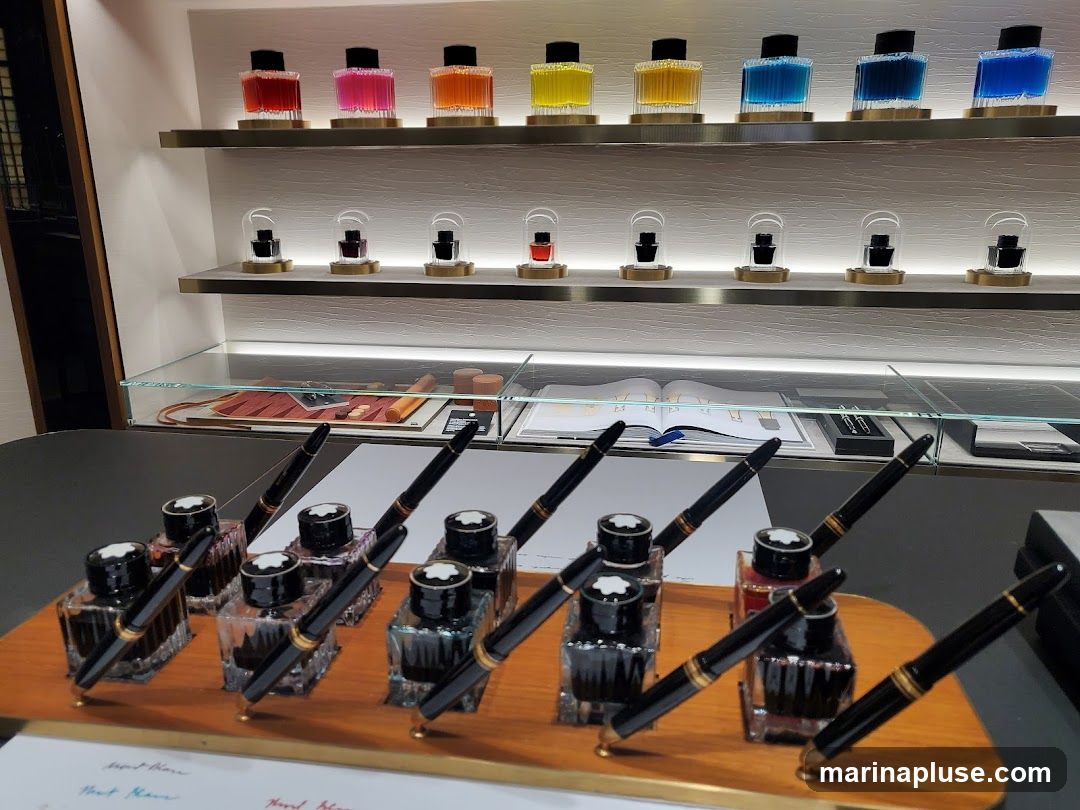 A display of Montblanc fountain pens and ink bottles is showcased on a wooden stand. The pens are black with gold accents, and each is placed in a small inkwell. Behind the pens, on shelves, are rows of colorful ink bottles in various shades of red, pink, orange, yellow, blue, and teal. Some ink bottles are displayed under glass domes. A glass display case in the background contains writing instruments and a book.