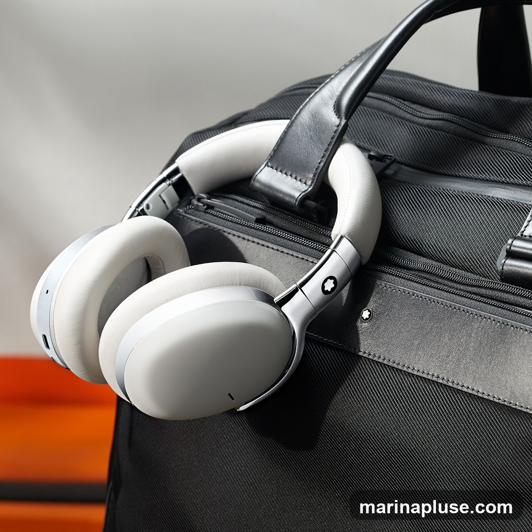 A pair of white Montblanc headphones rests on a black Montblanc bag. The headphones have silver accents and a white leather headband. The bag is made of textured black fabric with black leather handles and trim. A Montblanc star logo is visible on the headphones and the bag.