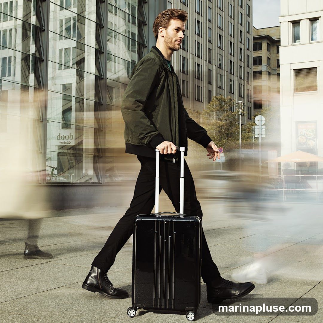 A man walks down a city street, pulling a black suitcase. He is wearing a dark green bomber jacket, black pants, and black boots. The background shows modern buildings and blurred figures of other pedestrians, suggesting movement and urban life.