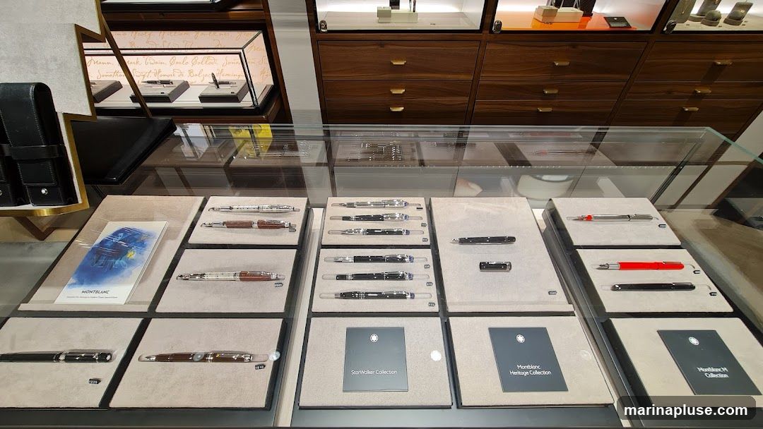 A display case filled with various Montblanc pens. The pens are arranged on white stands within the glass case. Some pens are silver with brown accents, others are black, and some are red. There are also display cards for the "StarWalker Collection" and "Montblanc Heritage Collection". In the background, wooden cabinets and shelves are visible, with more pens displayed on shelves above. A black leather pen case with the Montblanc logo is on the left side of the display.