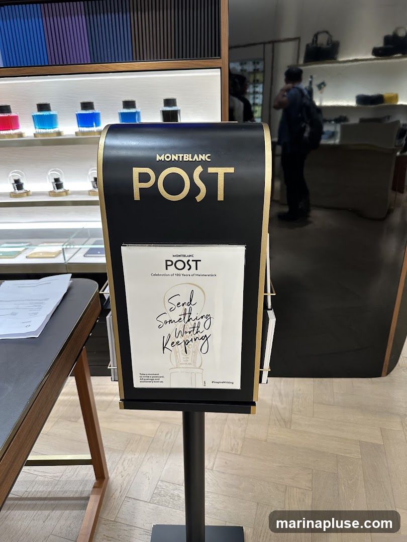 A black display stand with gold lettering that reads "MONTBLANC POST" is in the foreground. Below this, a brochure with the same branding and the slogan "Send Something Worth Keeping" is displayed. The background shows shelves with ink bottles and other Montblanc products, as well as a person browsing in the store.