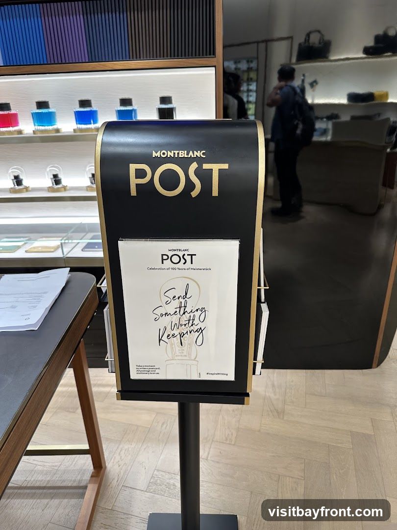 A black display stand with gold lettering that reads "MONTBLANC POST" is in the foreground. Below this, a brochure with the same branding and the slogan "Send Something Worth Keeping" is displayed. The background shows shelves with ink bottles and other Montblanc products, as well as a person browsing in the store.