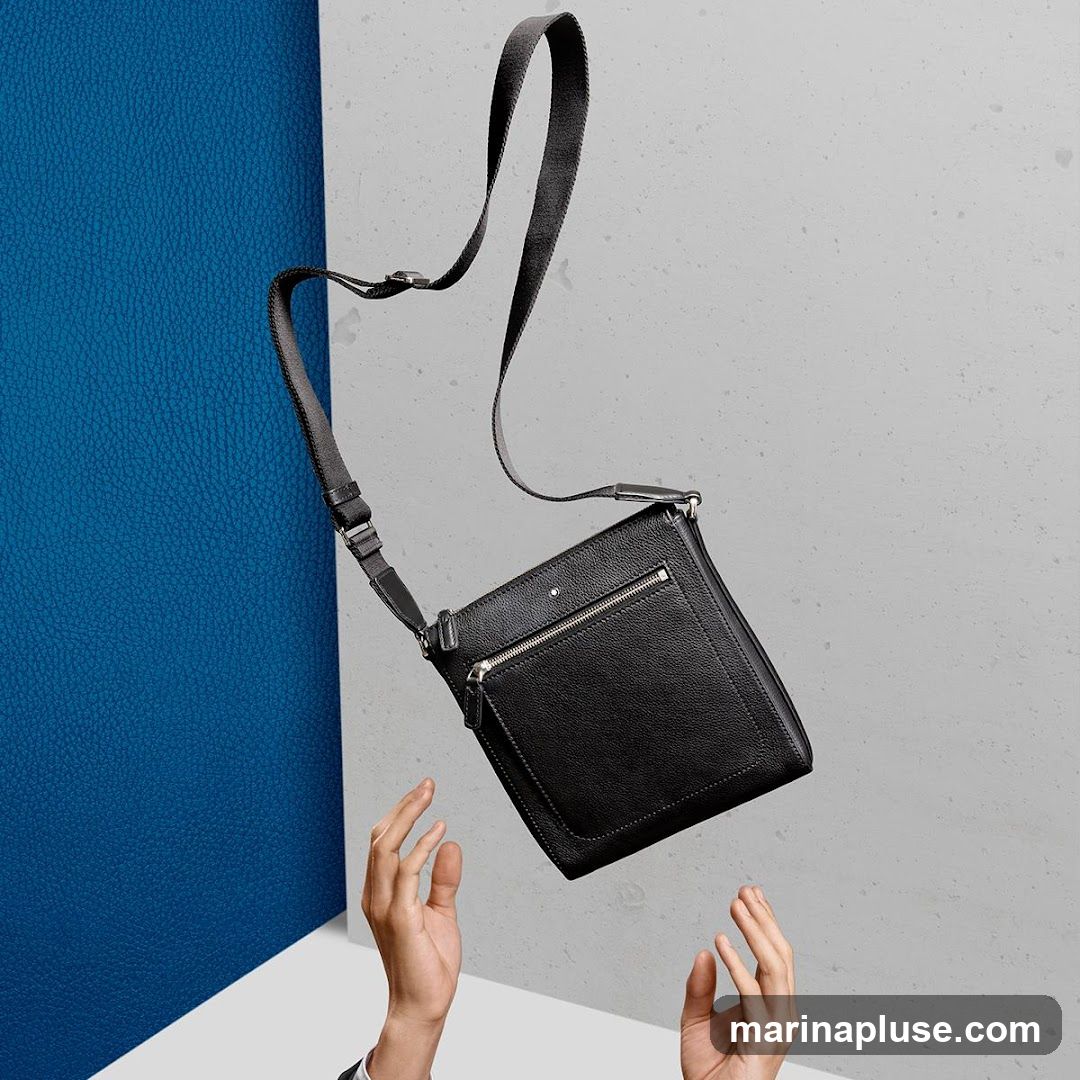 A black leather messenger bag with a zippered front pocket and a smaller pocket below it, is suspended in mid-air. A dark strap extends from the top of the bag. Two hands reach up towards the bag from the bottom of the frame. The background is split between a textured blue surface on the left and a speckled grey surface on the right.