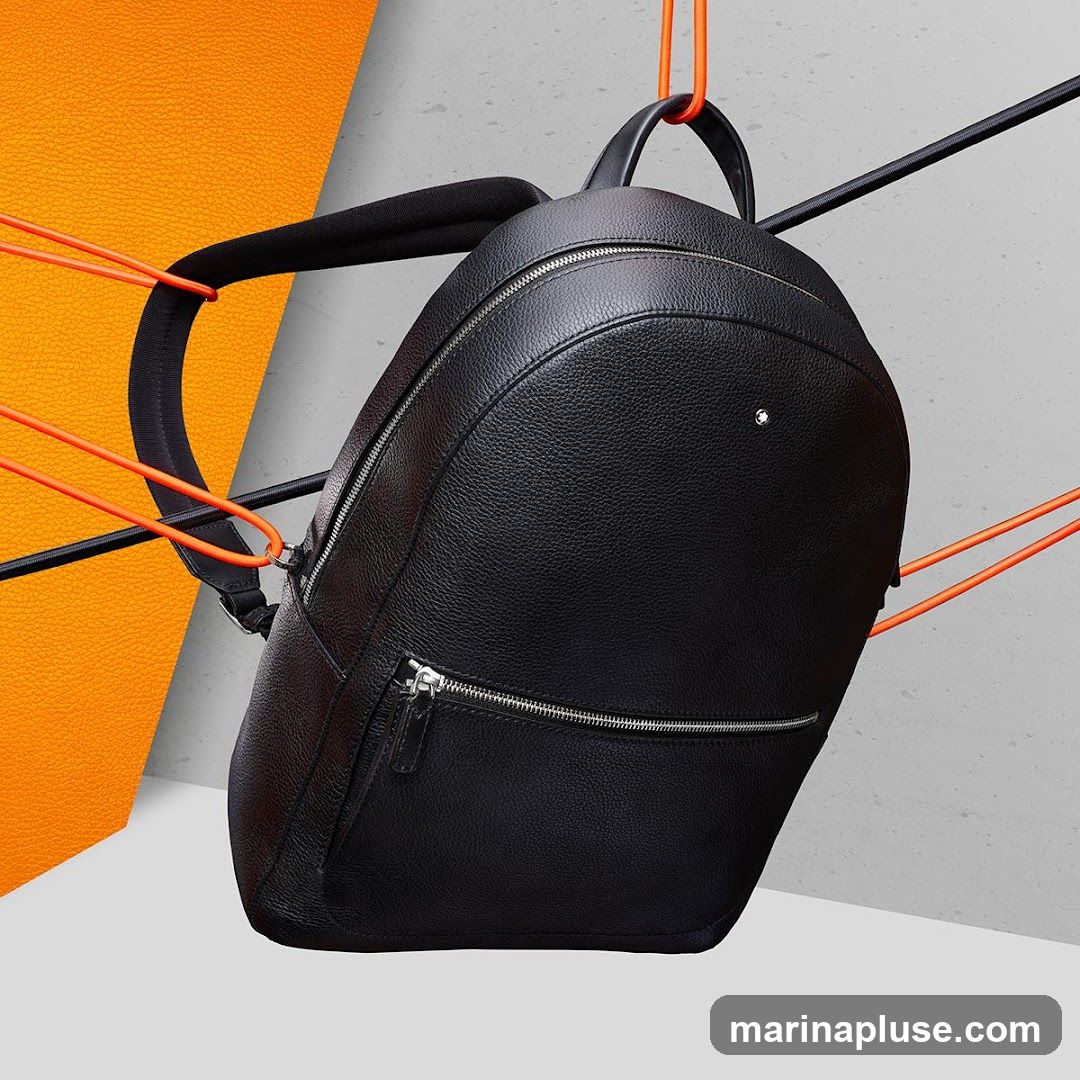 A black leather backpack with silver zippers is suspended by orange cords against a backdrop of textured orange and grey surfaces. The backpack has a small white emblem on its front.