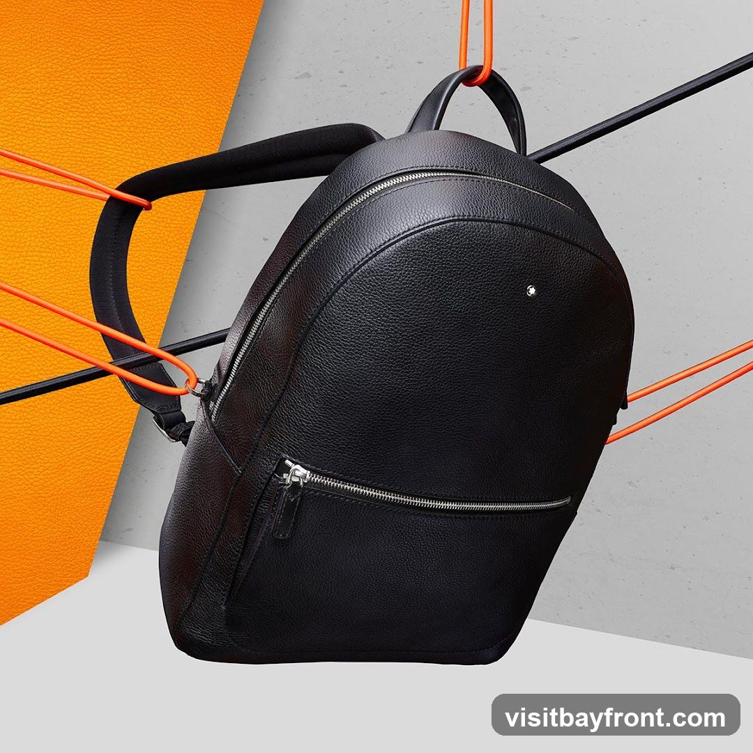 A black leather backpack with silver zippers is suspended by orange cords against a backdrop of textured orange and grey surfaces. The backpack has a small white emblem on its front.
