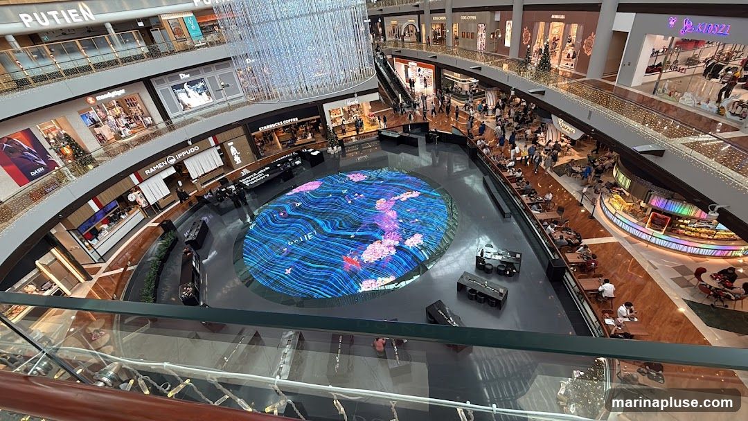 An aerial view of a bustling indoor shopping mall atrium. A large, oval-shaped LED screen in the center displays a vibrant, abstract design with pink floral elements and blue wavy lines. Surrounding the screen are various shops and dining establishments on multiple levels, with escalators and walkways connecting them. People are seen browsing, dining, and walking through the mall. Decorative lights adorn the railings and ceilings, adding to the festive atmosphere.