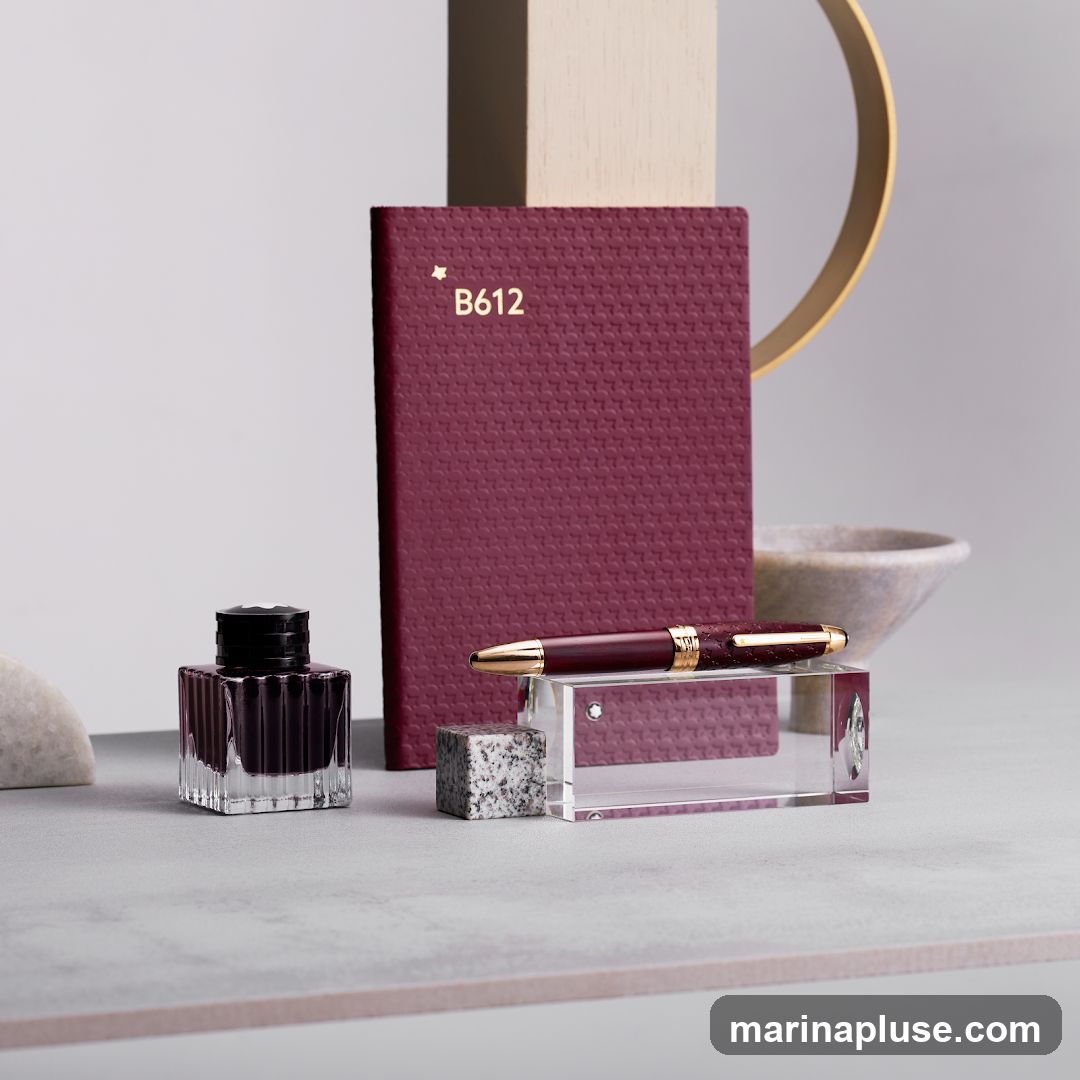 A burgundy notebook with gold lettering 'B612' and a star emblem is displayed next to a matching burgundy pen with gold accents. The pen rests on a clear acrylic stand. A glass inkwell filled with dark ink is positioned to the left of the notebook. The arrangement is set against a neutral background with abstract decorative elements, including a beige rectangular prism and a gold ring.