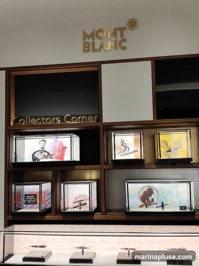 A display case featuring Mont Blanc pens and artwork. The Mont Blanc logo and "Collectors Corner" are visible. Several glass cases showcase pens and artistic prints, some with labels like "SOLD OUT" and "New". Below, a display case shows several pens on stands, with a background featuring handwritten names.