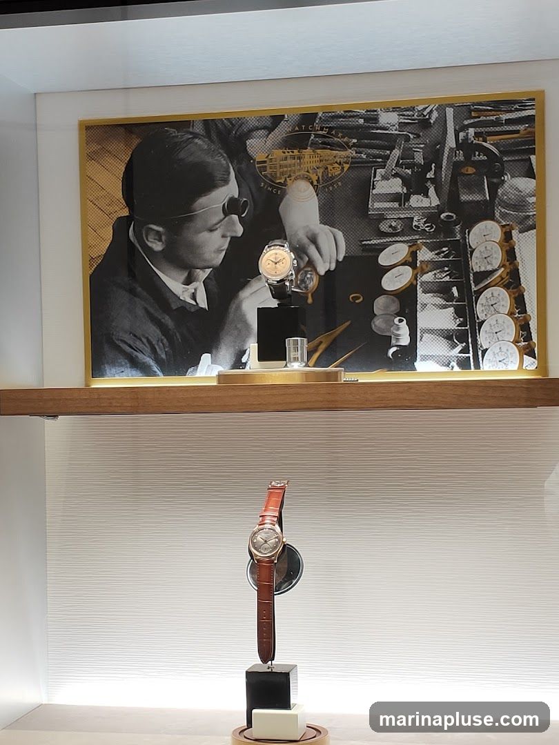 A display case featuring a vintage-style photograph of a watchmaker at work, with a modern wristwatch presented on a stand below. The photograph shows a man wearing a loupe, meticulously assembling a watch. The display also includes various watch components and tools. The wristwatch on display has a brown leather strap and a metallic face.