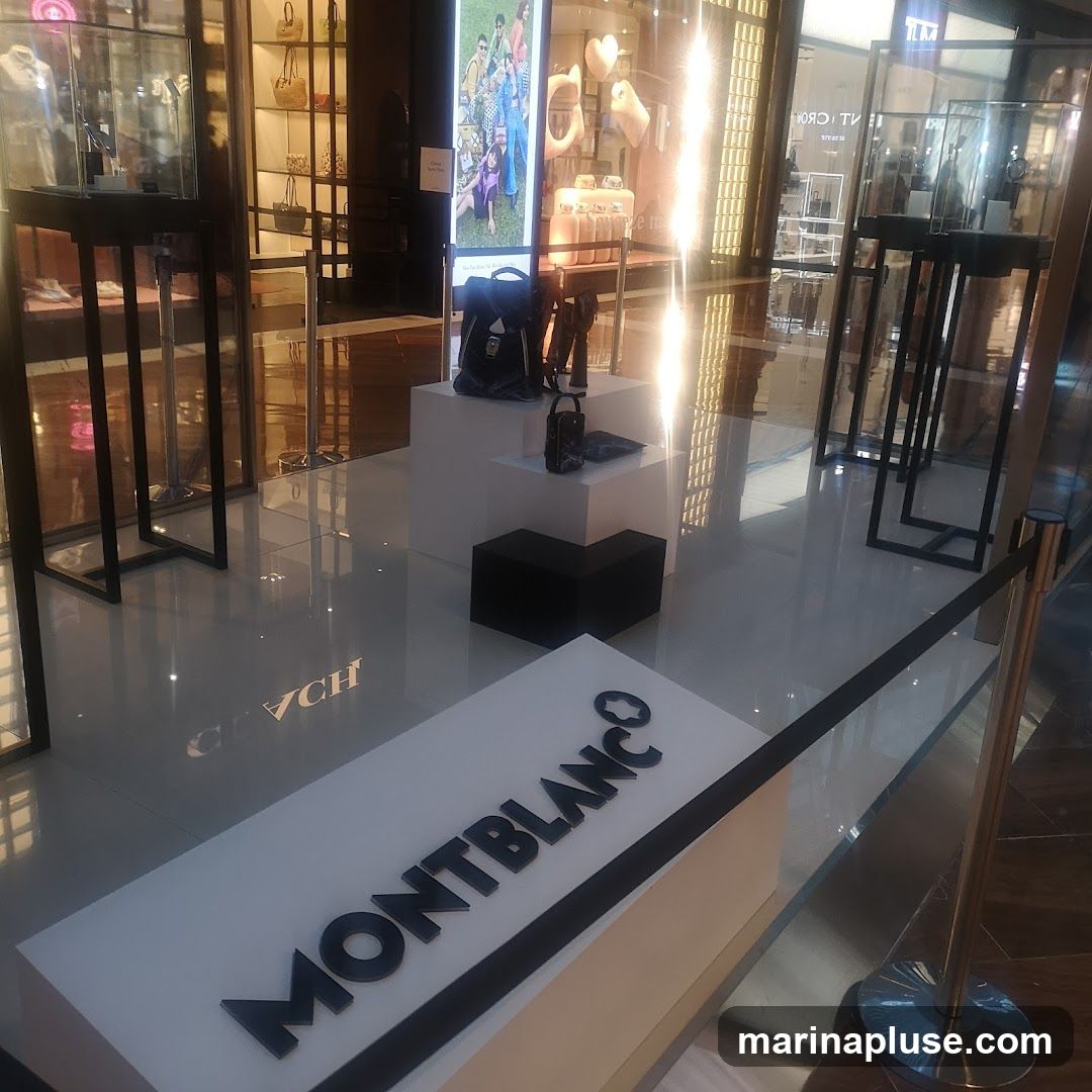A display of Montblanc merchandise, including bags and accessories, is set up in a retail environment. The Montblanc logo is prominently featured on a white platform in the foreground. In the background, there are glass display cases and illuminated panels showcasing other products.