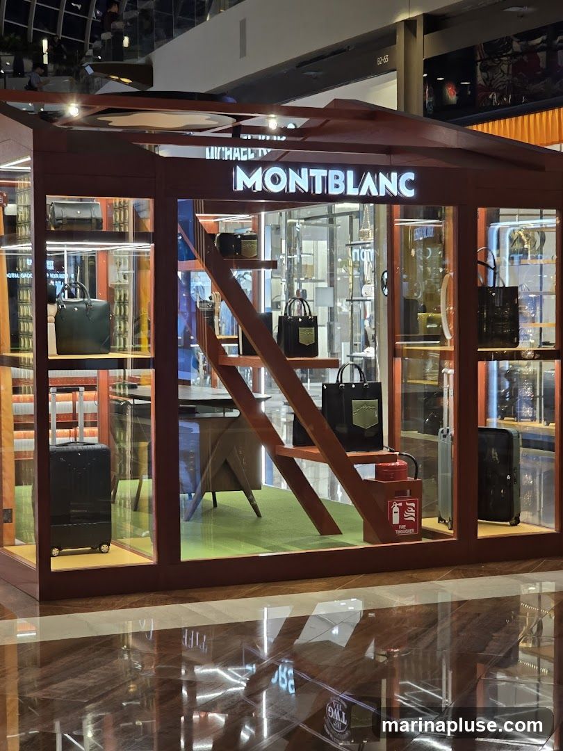 A display booth for Montblanc products, featuring handbags, luggage, and accessories. The booth has a modern design with wooden accents and glass panels. The Montblanc logo is prominently displayed above the entrance.
