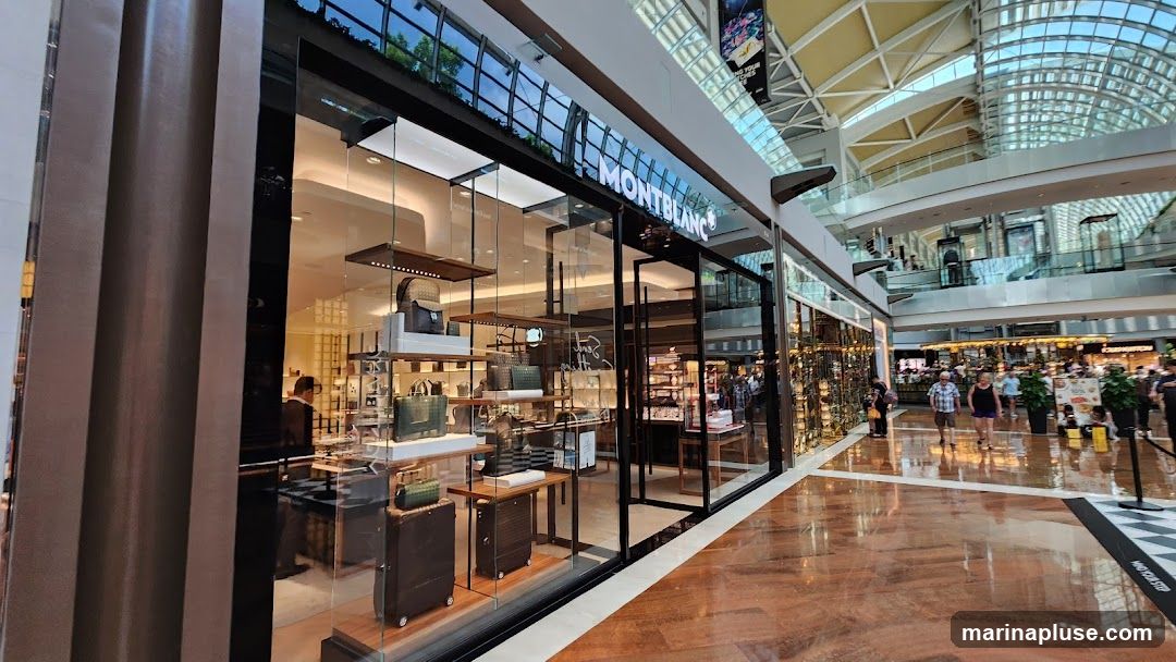 A wide shot of the Montblanc store's exterior in a modern shopping mall. The storefront features large glass windows displaying various luxury goods, including handbags, luggage, and accessories. The Montblanc logo is prominently displayed in illuminated white lettering above the entrance. Inside, shelves are neatly organized with products, and a sales associate can be seen assisting a customer. The mall's interior, with its high ceilings and architectural details, is visible in the background.