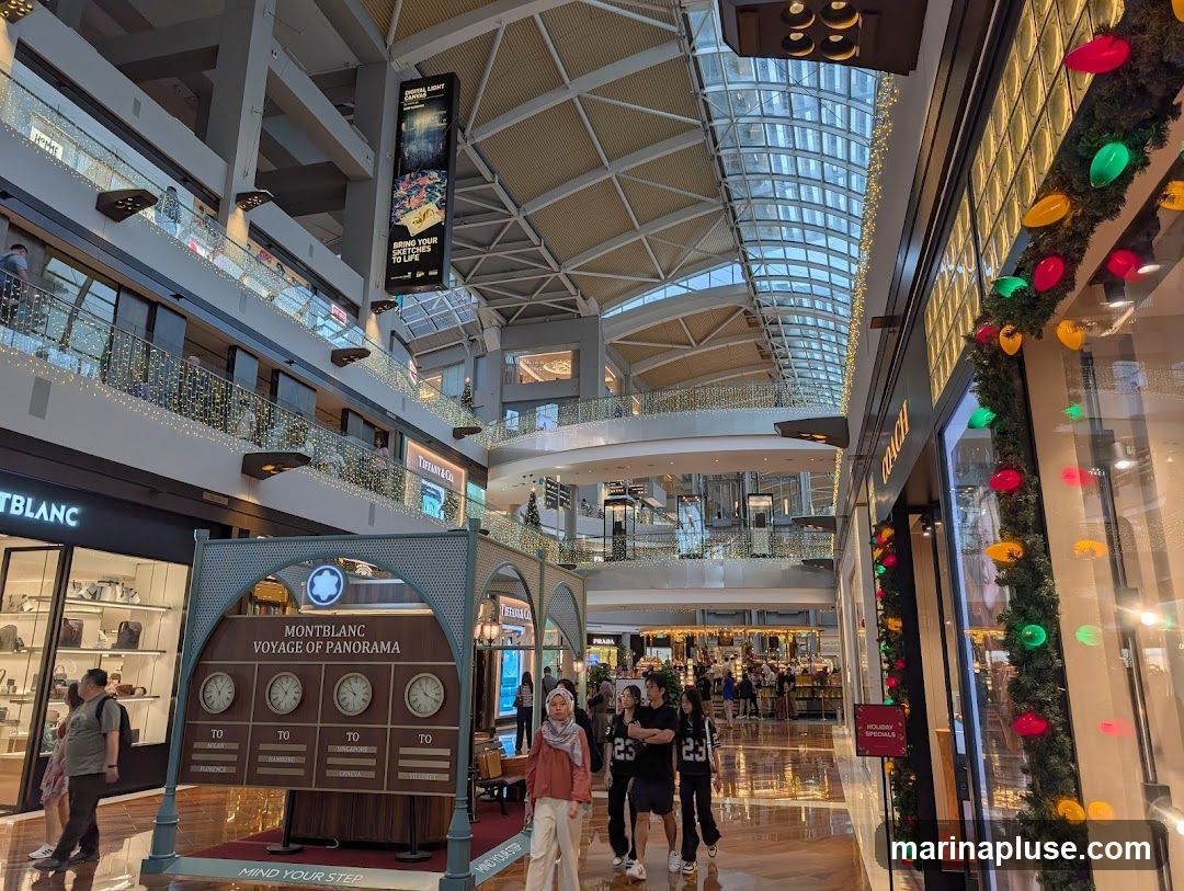 An interior view of a multi-level shopping mall with Christmas decorations. In the foreground, a Montblanc display features clocks showing times in different cities and the text "MONTBLANC VOYAGE OF PANORAMA". To the left is the Montblanc store. To the right, a store with festive lights and a "HOLIDAY SPECIALS" sign is visible. Other stores like Tiffany & Co. are also seen on different levels.