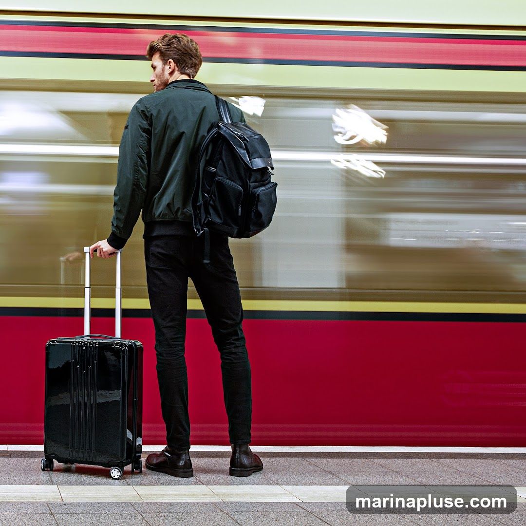 A man stands on a train platform, holding the handle of a rolling suitcase. He is wearing a dark green bomber jacket, black pants, and brown shoes. A black backpack is slung over his shoulder. A train is passing by in the background, blurred due to motion.