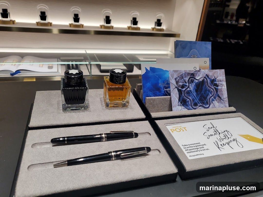 A display of Montblanc writing instruments and stationery. Two bottles of ink, black and amber, are shown alongside two pens. Several postcards with abstract designs are also displayed, along with a sign that reads "Montblanc POST. Send Something Worth Keeping. Take a moment to write a postcard. All postage and stationery is on us. #inspireWriting". In the background, shelves display watches and other items.