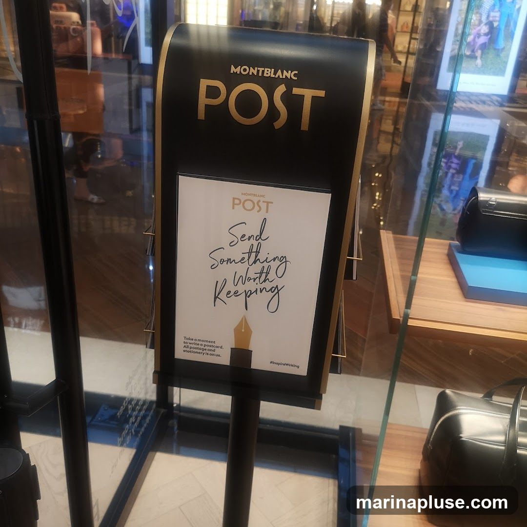 A black display stand with gold trim features the Montblanc logo and the word "POST" in large gold letters. Below this, a white card with the Montblanc Post logo reads "Send Something Worth Keeping." Smaller text at the bottom says, "Take a moment to write a postcard. All postage and stationery is on us." The hashtag "#inspireWriting" is also visible. The display is set up in front of a glass window, with merchandise visible inside the store.