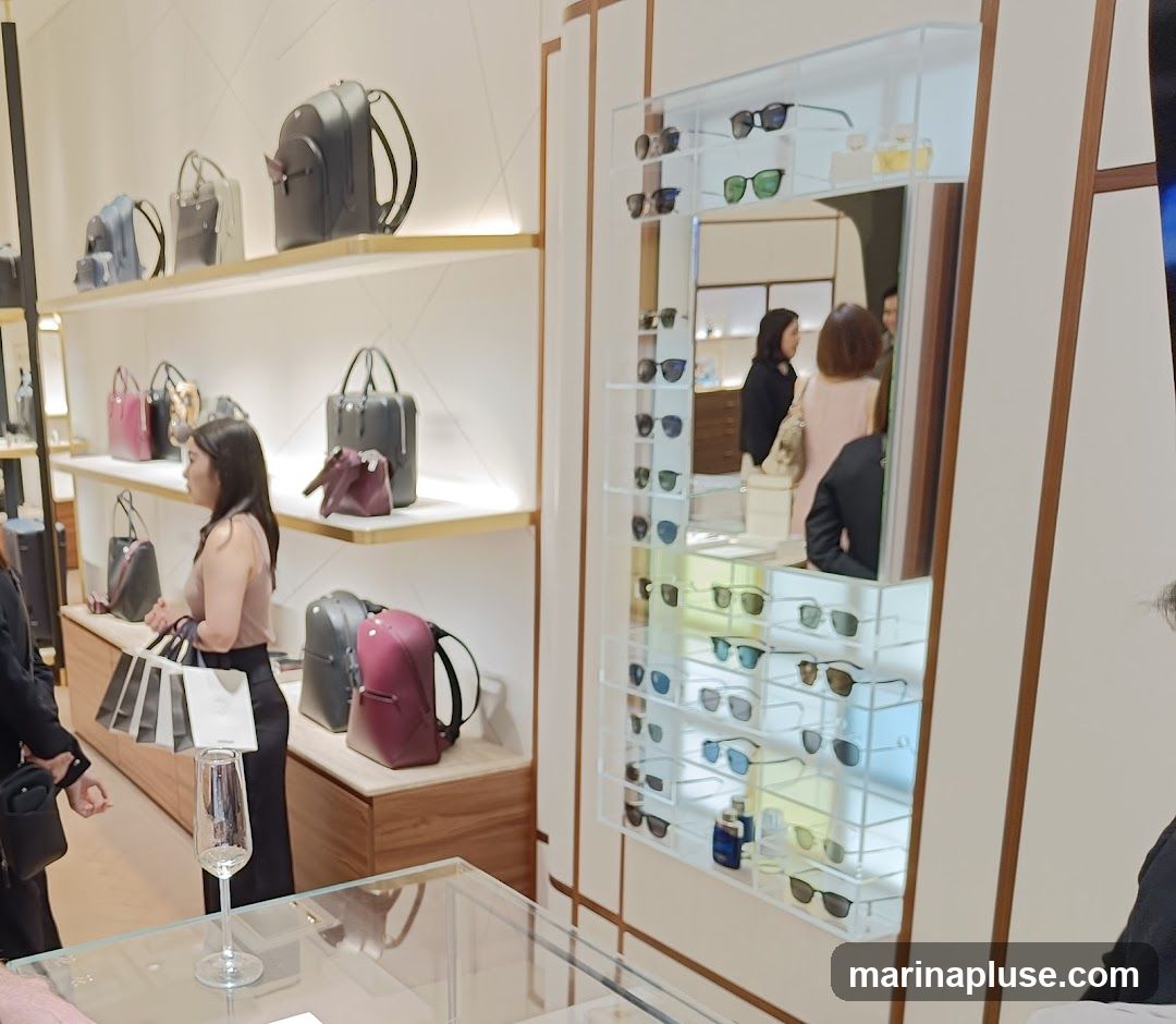 A retail store interior with shelves displaying various handbags and a vertical display case filled with sunglasses. A woman is seen browsing the handbags.