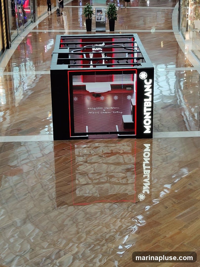 A Montblanc branded display kiosk is shown in a shopping mall. The kiosk is black with red accents and features the Montblanc logo and name. Inside the kiosk, there are shelves and displays, with a sign that reads "#My1810 Montblanc x (RED) Carbon Trolley". The floor is highly reflective, showing the reflection of the kiosk and the surrounding mall environment.