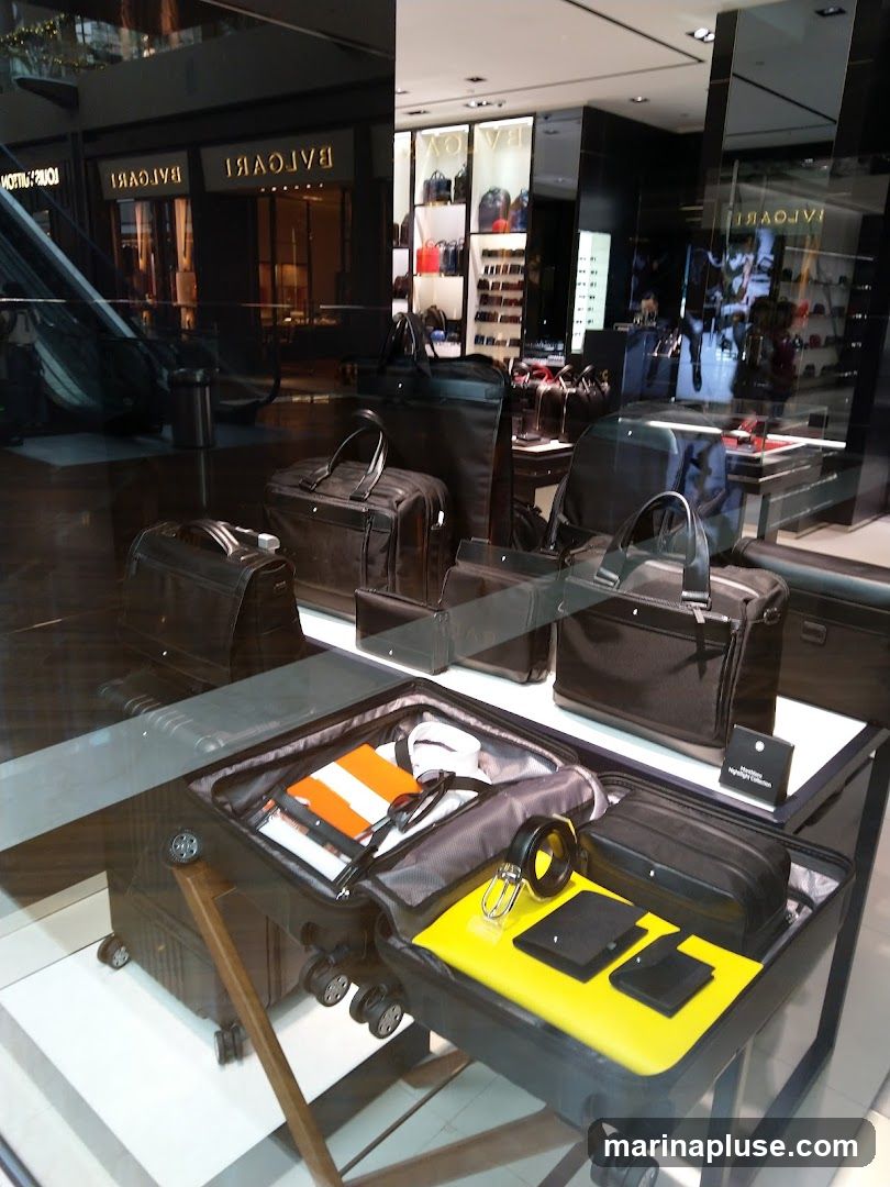 A display window showcases various luxury travel items, including open suitcases filled with neatly packed clothing, sunglasses, and accessories. Several leather bags and briefcases are also displayed on shelves and stands. The store's signage, "BULGARI," is visible in the background, reflected in the glass.