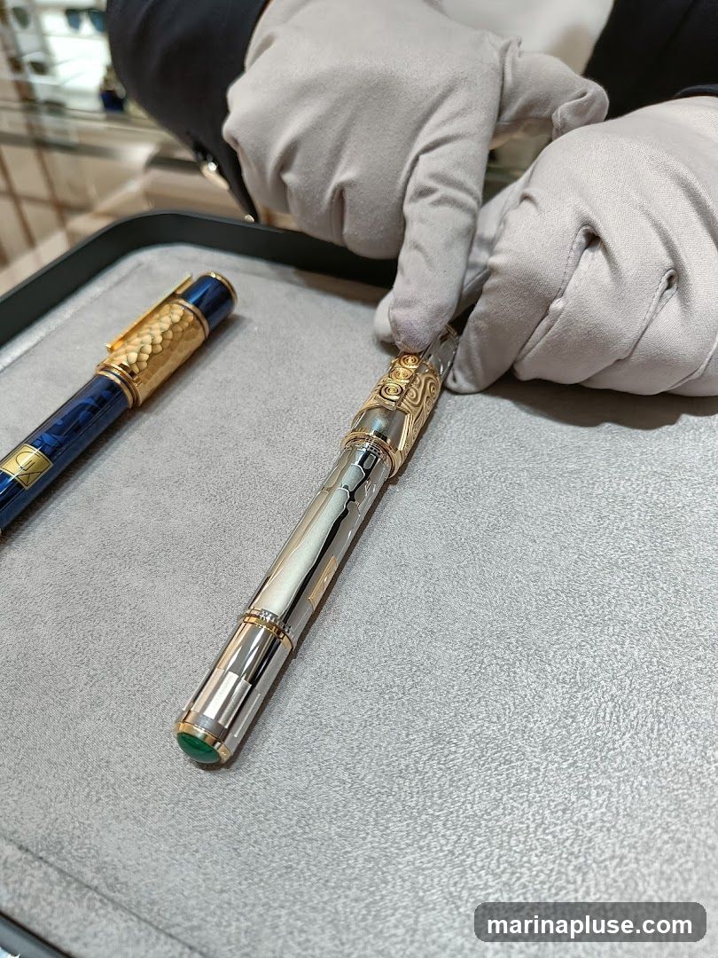 A close-up shot of two ornate pens being presented by gloved hands. The pen in the foreground is silver with gold accents and a green jewel at the end. The pen in the background is dark blue with a textured gold cap. The pens are displayed on a grey surface, possibly in a retail setting.