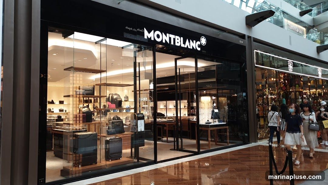 The exterior of a Montblanc store with a large glass facade. Inside, shelves display various leather goods, luggage, and accessories. The Montblanc logo is prominently displayed above the entrance. People are walking past the store.