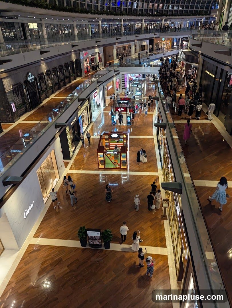 An overhead view of a multi-level shopping mall with many stores and shoppers. The mall features polished wooden floors and a modern architectural design with glass railings and extensive lighting. Various shops are visible, including Cartier, Coach, and Montblanc, along with display stands and promotional materials. People are seen browsing, walking, and taking photos.