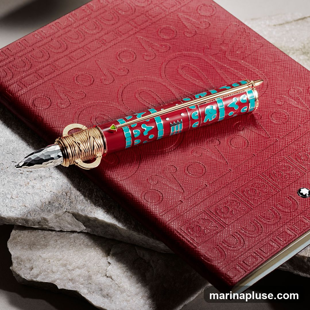 A close-up shot of a red notebook with embossed patterns and a Montblanc logo. A decorative pen with red and turquoise accents and gold wire detailing rests on top of the notebook. The notebook is placed on rough, light-colored stones.
