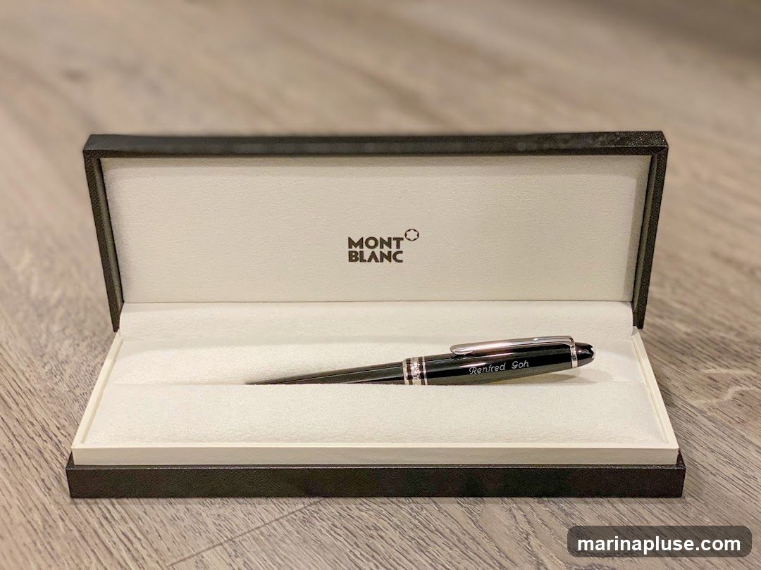 A black Montblanc pen with silver accents is displayed in its open black and white presentation box. The pen has the name "Renfred Soh" engraved on it. The Montblanc logo is visible on the inside lid of the box. The box is placed on a wooden surface.