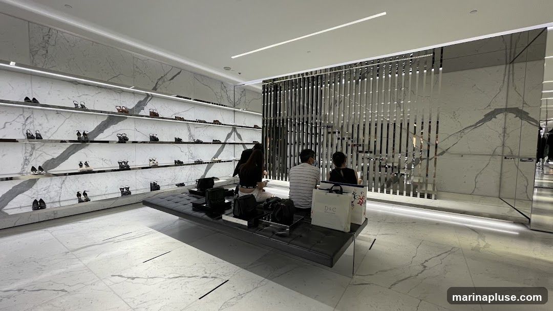 A modern and luxurious shoe store interior with marble-patterned walls and floors. Shelves display a variety of high-heeled shoes. A long, dark bench in the foreground is occupied by three people, two women and one man, who appear to be resting or browsing. Several shopping bags are placed on the bench. A decorative metal screen separates the seating area from a staircase in the background.