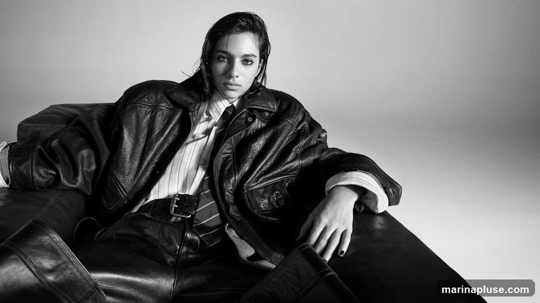 A black and white studio portrait of a person wearing a leather jacket, a striped shirt, and a tie, seated on a dark surface. The person has wet hair and is looking directly at the camera.