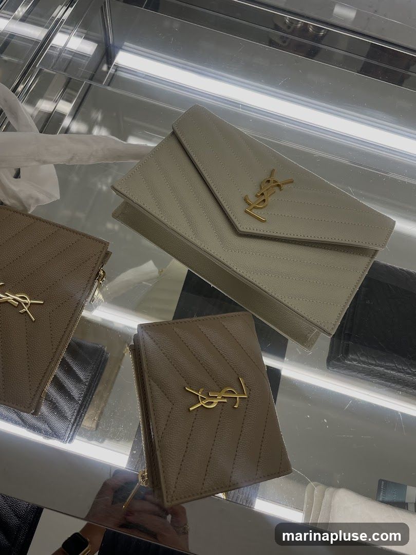 A display of designer wallets and a clutch bag with the YSL logo, presented in a glass display case with interior lighting. The items are in neutral tones of beige and white, with gold-tone hardware.