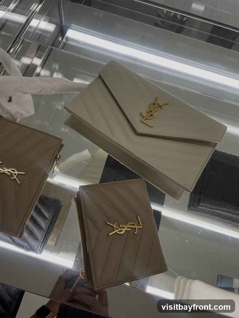 A display of designer wallets and a clutch bag with the YSL logo, presented in a glass display case with interior lighting. The items are in neutral tones of beige and white, with gold-tone hardware.