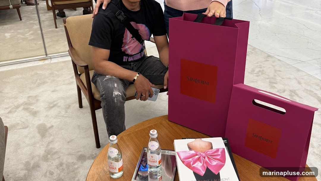A person is sitting on a chair next to several magenta shopping bags with the Saint Laurent logo. Two bottles of Acqua Panna water and a book with a large pink bow on the cover are on a wooden table.