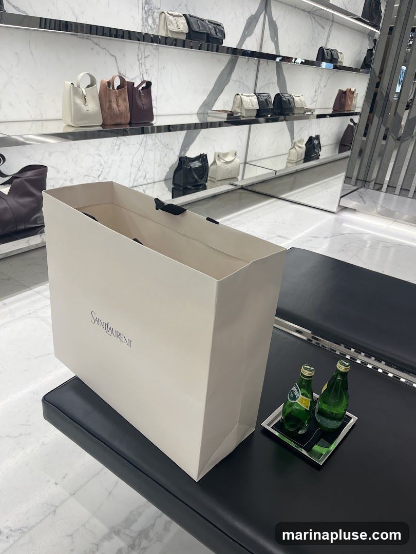 A white shopping bag with the Saint Laurent logo is placed on a black bench. Next to the bag are two green glass bottles of Perrier water on a silver tray. In the background, shelves display various handbags.