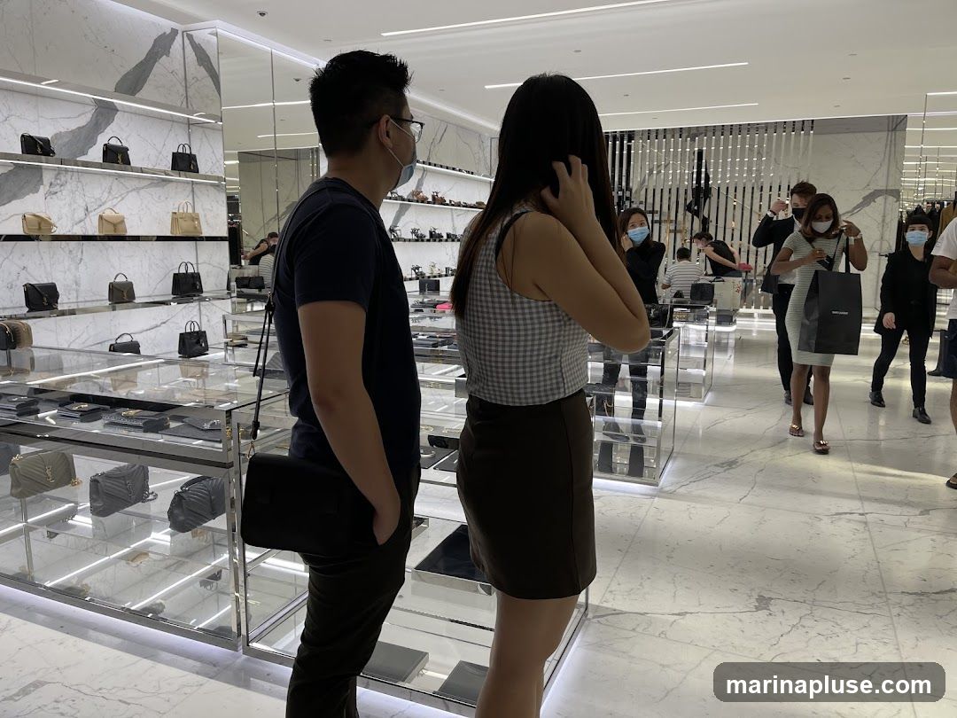 People browsing handbags and accessories in a brightly lit, modern luxury store. Displays feature various handbags on shelves and in glass cases. Several customers, some wearing masks, are seen looking at merchandise. One woman is holding a shopping bag with a visible logo.