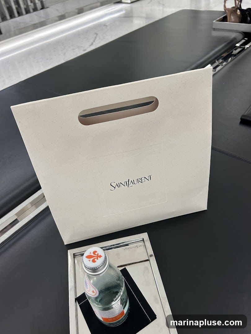 A white shopping bag with the Saint Laurent logo is placed on a black surface. Next to it, a glass bottle of water with an orange and white cap sits on a silver tray with a black napkin.