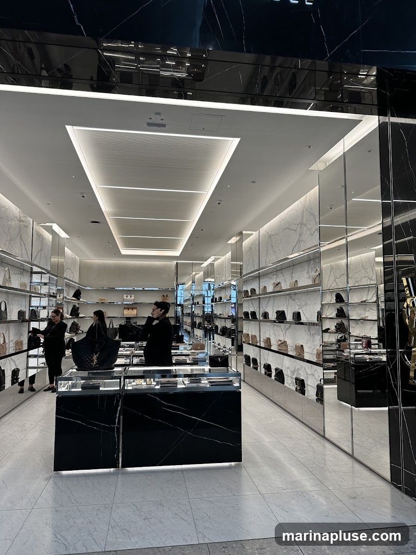 The interior of a high-end store, likely selling luxury handbags and accessories. The store features a sleek, modern design with white marble-patterned walls and shelves, mirrored accents, and black marble display cases. Numerous handbags are displayed on the shelves and in the display cases. Three individuals, presumably staff or customers, are visible within the store. The lighting is bright and modern, with recessed ceiling lights and strip lighting along the shelves.