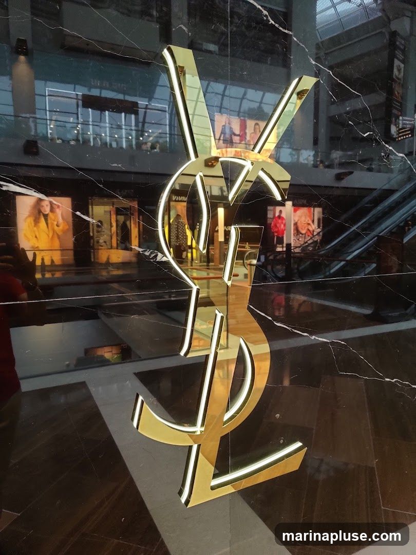 A large, illuminated gold YSL logo is prominently displayed on a black marble wall. Behind the logo, reflections show a shopping mall interior with escalators, storefronts, and people. Several fashion advertisements featuring models in colorful clothing are visible on the walls of the mall.