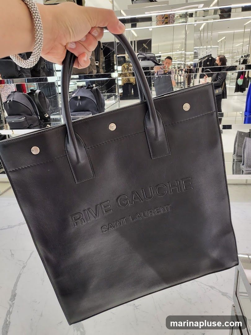 A person is holding a black leather tote bag with "RIVE GAUCHE SAINT LAURENT" embossed on it. The bag has two top handles and silver stud details. In the background, a retail store interior is visible with clothing racks, mirrors, and other shoppers.