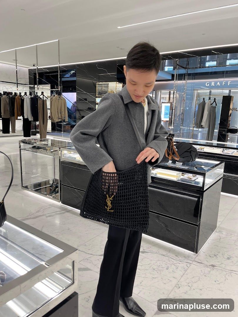 A person in a grey coat and black pants holds a black mesh tote bag with a gold Yves Saint Laurent logo. The person is standing in a brightly lit store with displays of clothing and accessories.